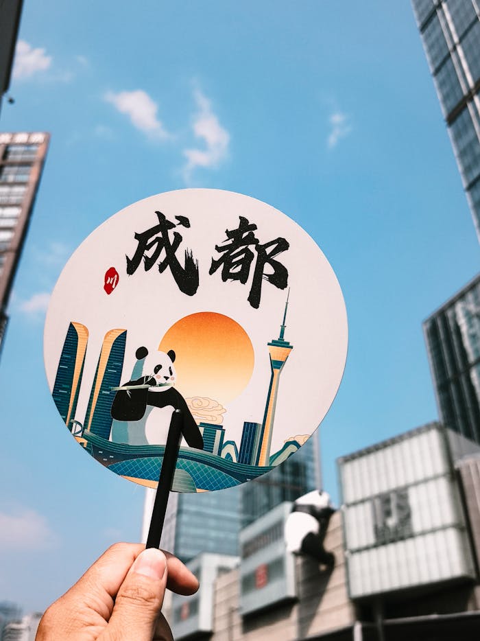 Person holding a decorative Chengdu fan with a city skyscraper backdrop on a sunny day.